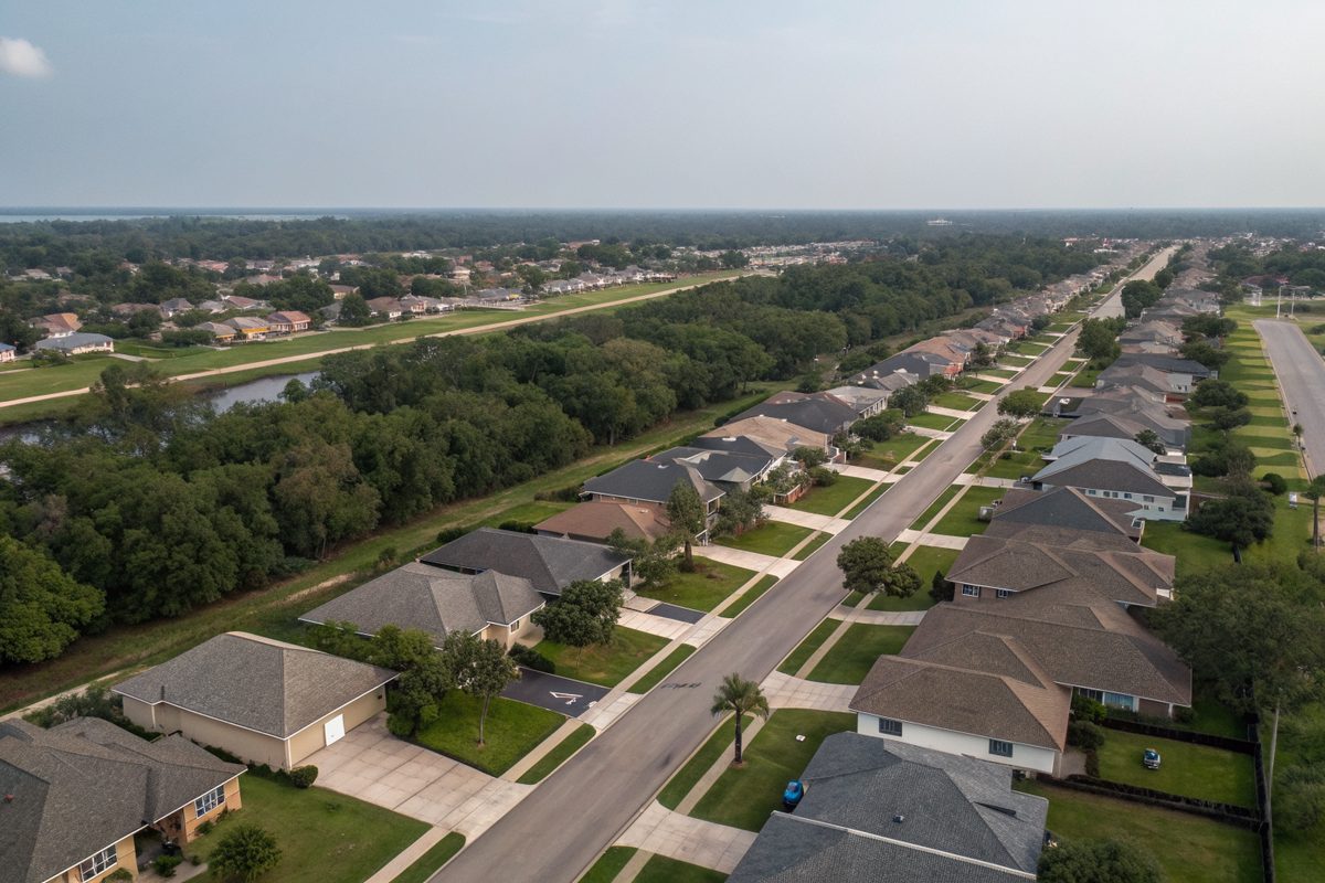 Aerial view of a flat neighborhood of shingle roofs, live oaks and a drainage channel