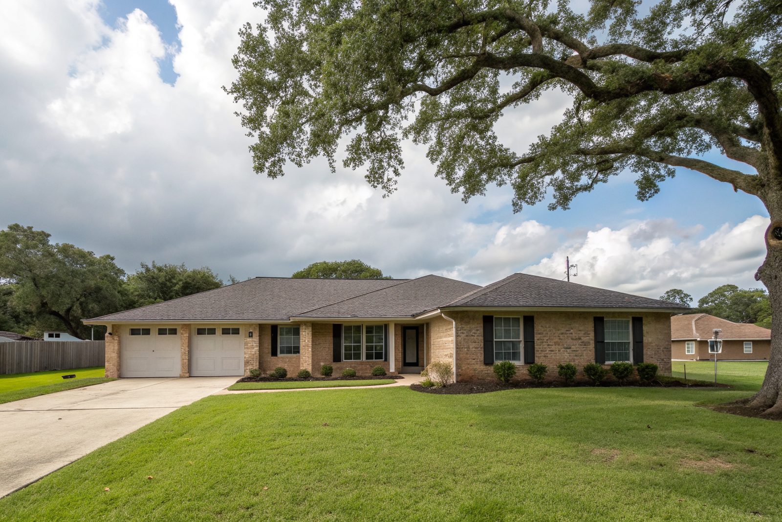 A one-story tan brick ranch home with a charcoal shingle roof and a large live oak in the yard