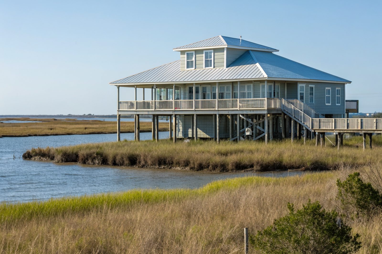 A raised bay house on pilings with a light gray standing-seam metal roof beside marsh grass